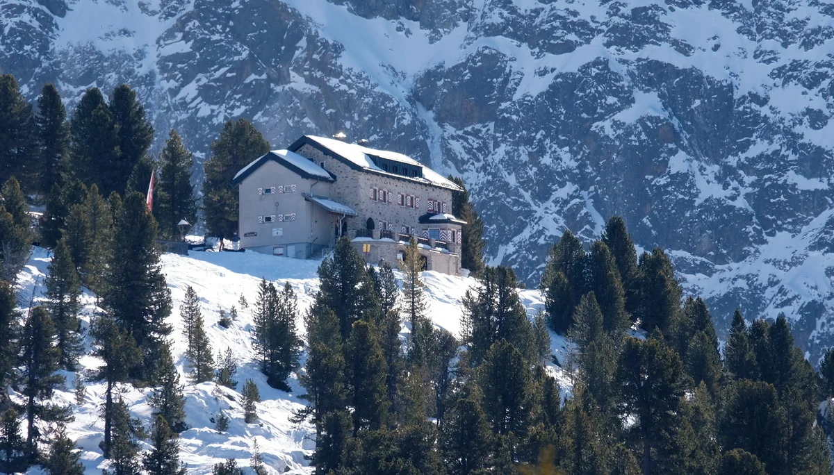 Bielefelder Hütte im Winter. Die Hütte liegt auf einem Hügel, der mit Schnee bedeckt und von der Sonne beschienen ist. Rings herum steht Tannen und Fichten, die grün sind, also keine Schneeauflagen haben. Hinter dem Hügel mit der Hütte sieht man die steil emporragende, teils mit Schnee bedeckte Felswand.  Vor der Hütte befindet sich eine Panoramaterrasse, die nach Westen ausgeichtetist. An den hölzernen Fensterläden erahnt man die im Bielefelder Wappen vorkommende,  weiß-rote, schräg nach oben gerichtete, in der Mitte zulaufende Balken. AUf dem Dach liegt Schnee | © DAV Bielefeld