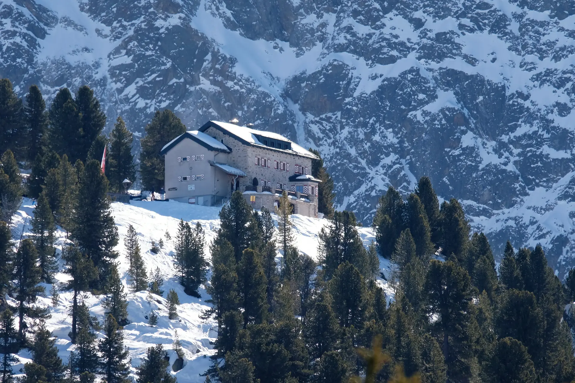 Bielefelder Hütte im Winter. Die Hütte liegt auf einem Hügel, der mit Schnee bedeckt und von der Sonne beschienen ist. Rings herum steht Tannen und Fichten, die grün sind, also keine Schneeauflagen haben. Hinter dem Hügel mit der Hütte sieht man die steil emporragende, teils mit Schnee bedeckte Felswand.  Vor der Hütte befindet sich eine Panoramaterrasse, die nach Westen ausgeichtetist. An den hölzernen Fensterläden erahnt man die im Bielefelder Wappen vorkommende,  weiß-rote, schräg nach oben gerichtete, in der Mitte zulaufende Balken. AUf dem Dach liegt Schnee | © DAV Bielefeld