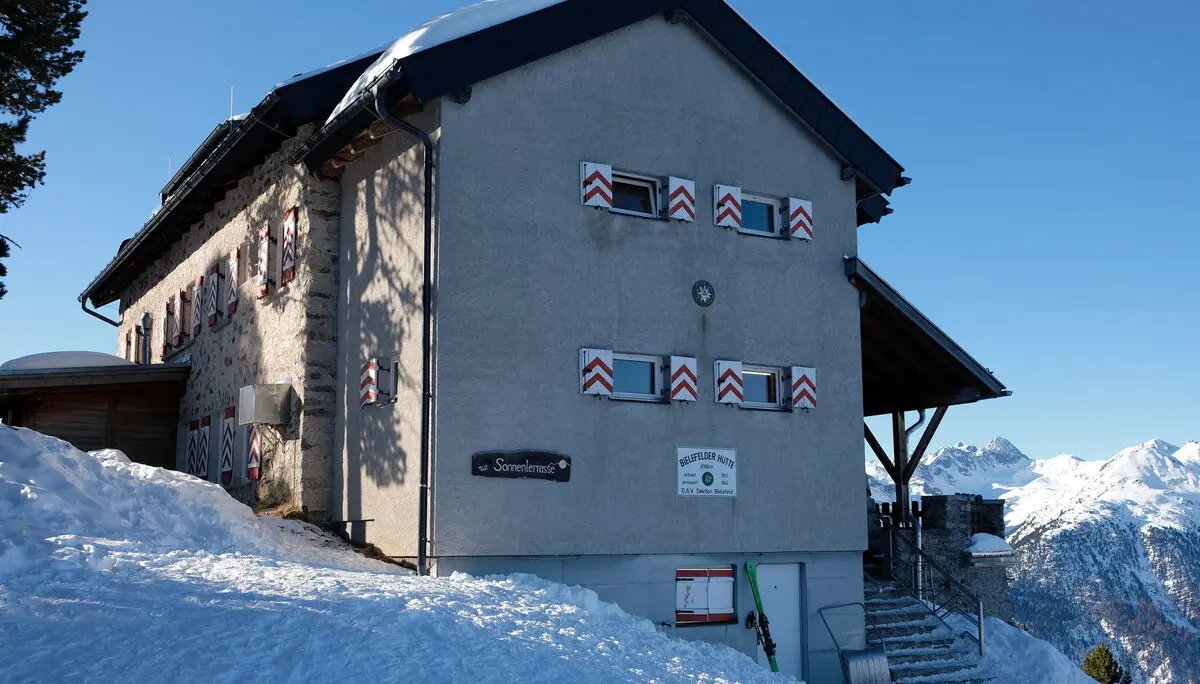 Die Bielefelder Hütte im Winter. Eine Seitenansicht von Norden, die einen grau verputzen Anbau mit dem Eingangsbereich zeigt. Im Vordergrund links liegt Schnee und es geht etwas bergauf. Rechts von der Hütte sieht man in der Ferne schneebedeckte Berge. Unter den vier Fenstern der Stirnseite der Hütte ist das weiße Schild mit der Aufschrift "Bielefelder Hütte " angebracht. Weiter liest man auf dem Schild: 2112m, erbaut1913, erneuert 1954, D.A.V. Sektion Bielefeld | © DAV Bielefeld
