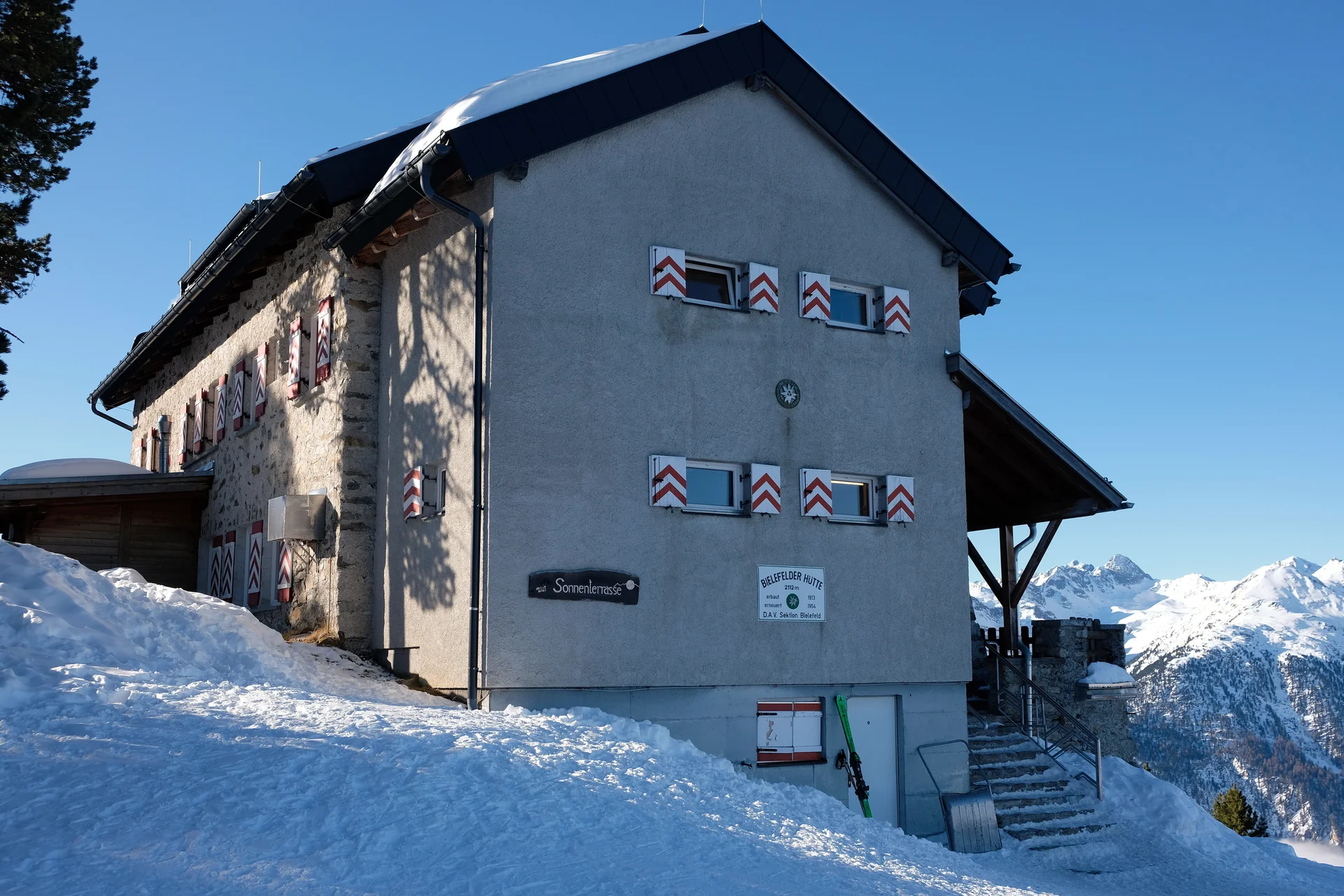 Die Bielefelder Hütte im Winter. Eine Seitenansicht von Norden, die einen grau verputzen Anbau mit dem Eingangsbereich zeigt. Im Vordergrund links liegt Schnee und es geht etwas bergauf. Rechts von der Hütte sieht man in der Ferne schneebedeckte Berge. Unter den vier Fenstern der Stirnseite der Hütte ist das weiße Schild mit der Aufschrift "Bielefelder Hütte " angebracht. Weiter liest man auf dem Schild: 2112m, erbaut1913, erneuert 1954, D.A.V. Sektion Bielefeld | © DAV Bielefeld