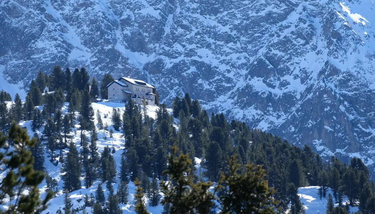 Bielefelder Hütte im Winter. Die Hütte liegt auf einem Hügel, der mit Schnee bedeckt und von der Sonne beschienen ist. Rings herum steht Tannen und Fichten, die grün sind, also keine Schneeauflagen haben. Hinter dem Hügel mit der Hütte sieht man die steil emporragende, teils mit Schnee bedeckte Felswand. Die BIelefelder Hütte sieht gegen die Felswand eher klein aus. | © DAV Bielefeld