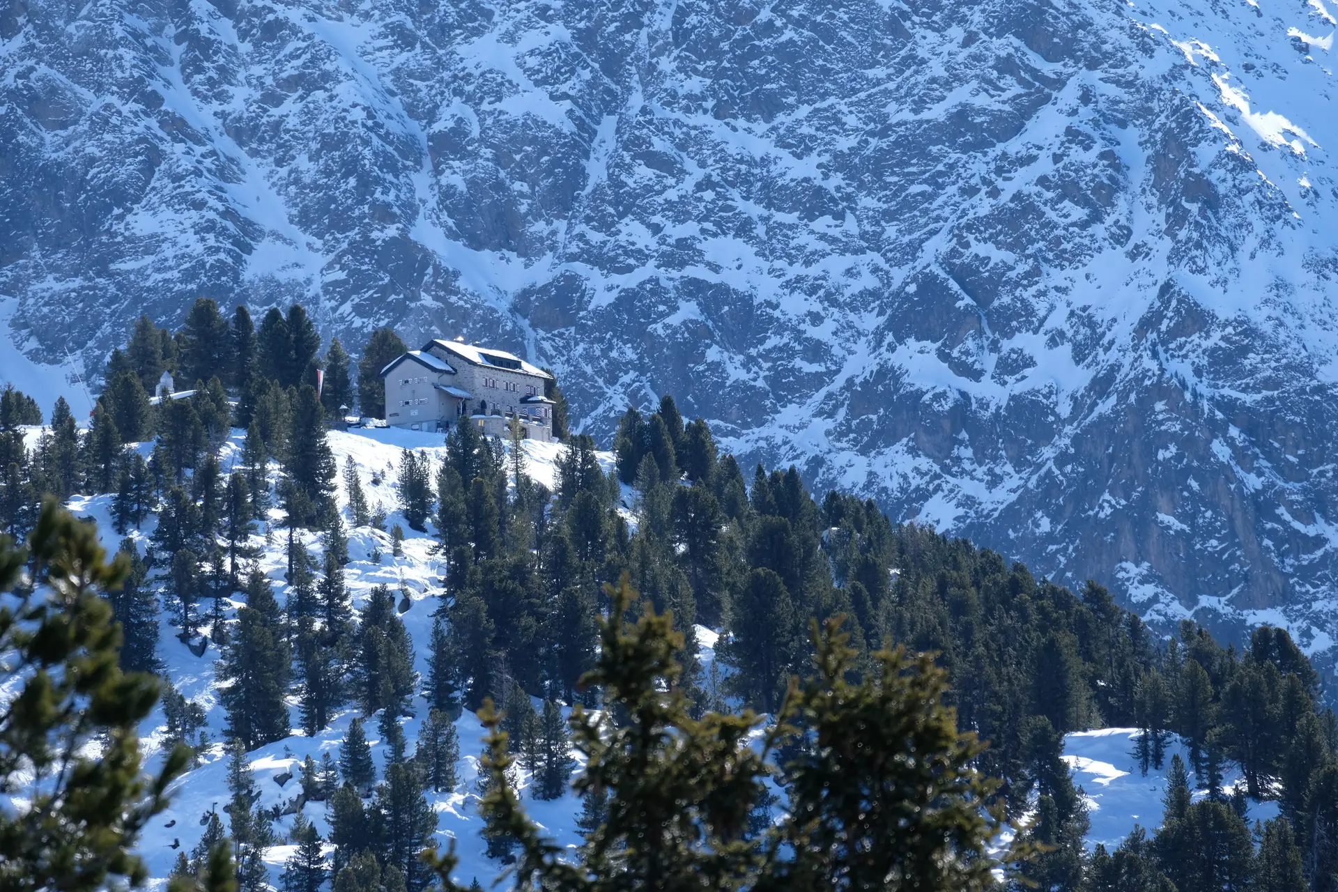 Bielefelder Hütte im Winter. Die Hütte liegt auf einem Hügel, der mit Schnee bedeckt und von der Sonne beschienen ist. Rings herum steht Tannen und Fichten, die grün sind, also keine Schneeauflagen haben. Hinter dem Hügel mit der Hütte sieht man die steil emporragende, teils mit Schnee bedeckte Felswand. Die BIelefelder Hütte sieht gegen die Felswand eher klein aus. | © DAV Bielefeld